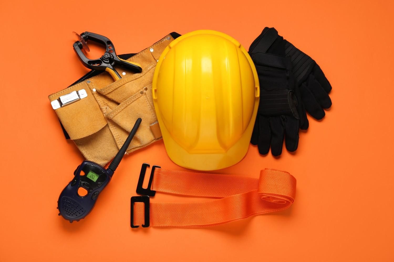 Casco amarillo, cinturón de herramientas, guantes, walkie-talkie y correa de seguridad naranja sobre fondo naranja.