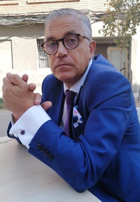 Hombre con traje azul, gafas y corbata morada, inclinado hacia delante, con las manos entrelazadas.