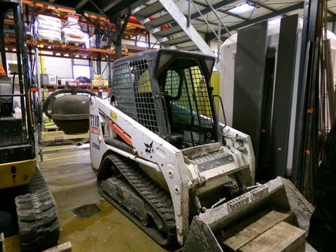 Chargeuse compacte blanche dans un entrepôt, godet levé et chenilles boueuses.