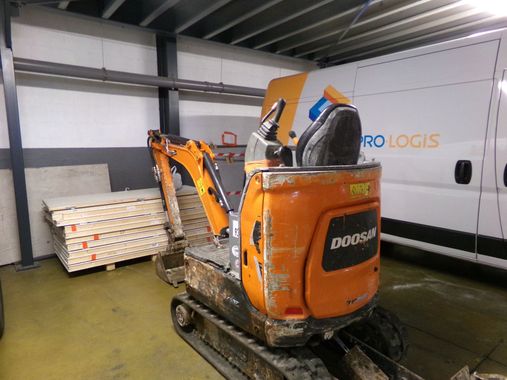 Une mini-pelle Doosan orange se trouve dans un entrepôt, à côté de palettes empilées et d'un camion Pro-Logis.