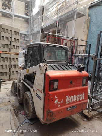 Chargeuse compacte Bobcat rouge dans une ruelle de chantier étroite, à côté de parpaings empilés et d'échafaudages.