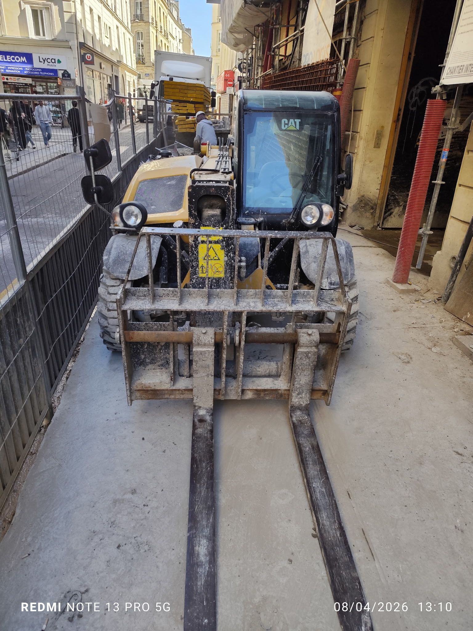 Un chariot élévateur, fourches levées, se trouve dans une rue étroite en centre-ville, à côté d'un trottoir et de bâtiments.