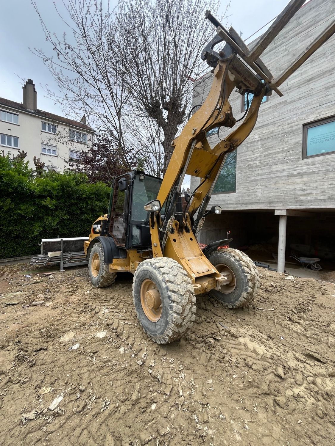 Une chargeuse articulée Caterpillar dans un jardin en friche, à côté d'une maison grise.