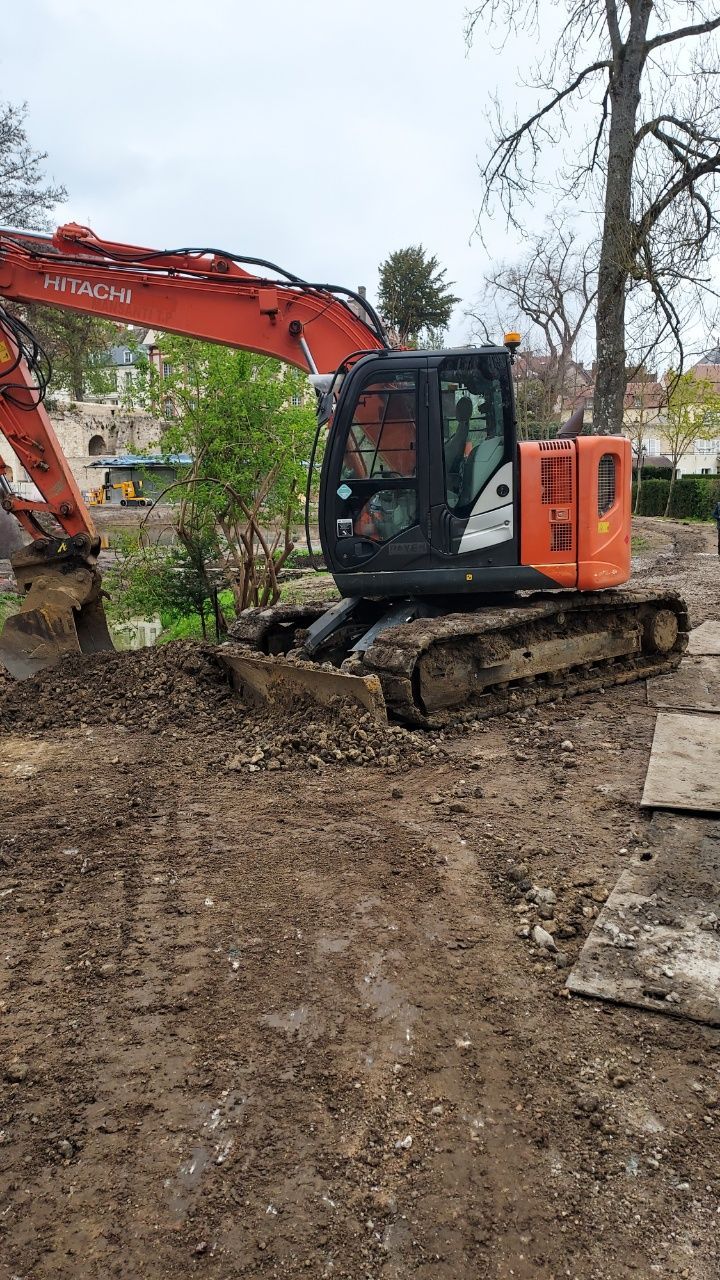 Une pelleteuse orange Hitachi travaille sur un terrain boueux, à côté d'arbres.
