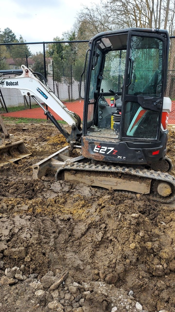 Une mini-pelle creuse dans un terrain boueux sur un chantier de construction derrière une clôture.