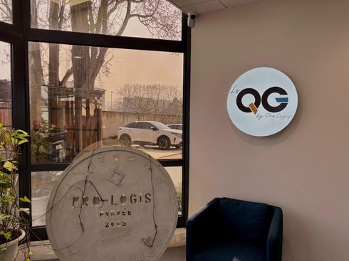 Hall d'entrée avec porte vitrée, logo mural Pro-Logis, chaise noire et panneau rond près d'une fenêtre.
