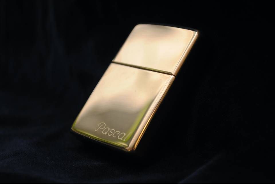 Ein goldfarbenes, rechteckiges Feuerzeug mit der eingravierten Aufschrift „Pascal“ vor dunklem Hintergrund.