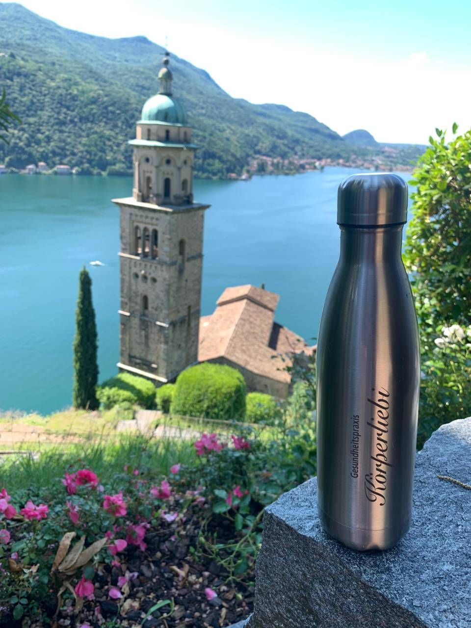 Edelstahl-Wasserflasche mit Inschrift, in der Nähe einer Steinkirche mit Blick auf einen See und Berge.