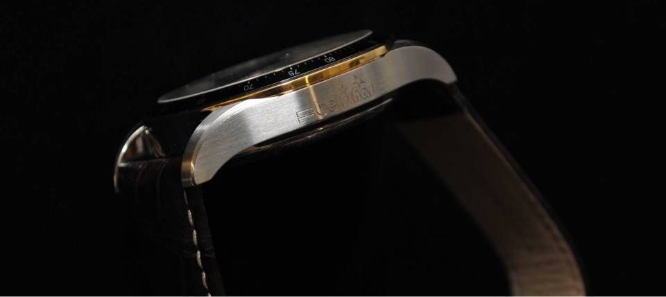 Nahaufnahme einer Armbanduhr in Seitenansicht, die ein Edelstahlgehäuse und ein schwarzes Armband vor schwarzem Hintergrund zeigt.