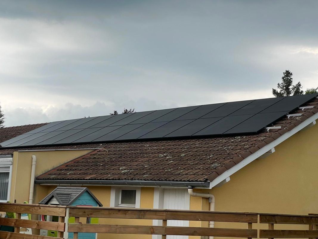 Panneaux solaires sur le toit de tuiles brunes d'une maison jaune, se détachant sur un ciel nuageux.