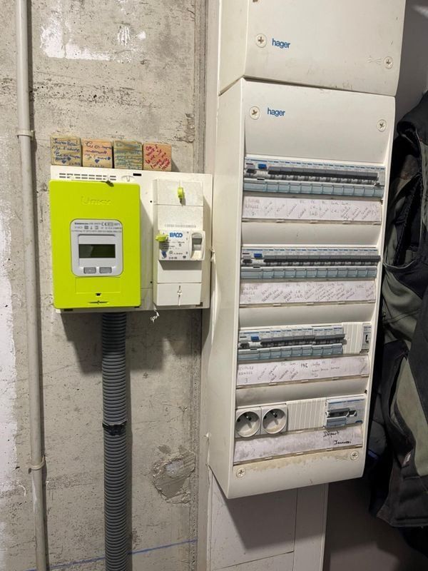 Tableau électrique avec compteur vert, boîtes blanches et disjoncteurs contre un mur en béton.