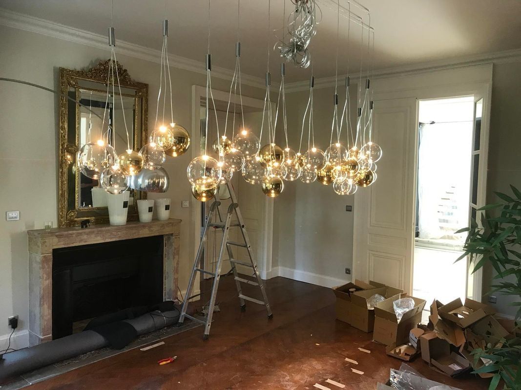 Pièce luxueuse avec des lustres ornés en cours d'installation près d'une cheminée. Une échelle et des caisses sont présentes.