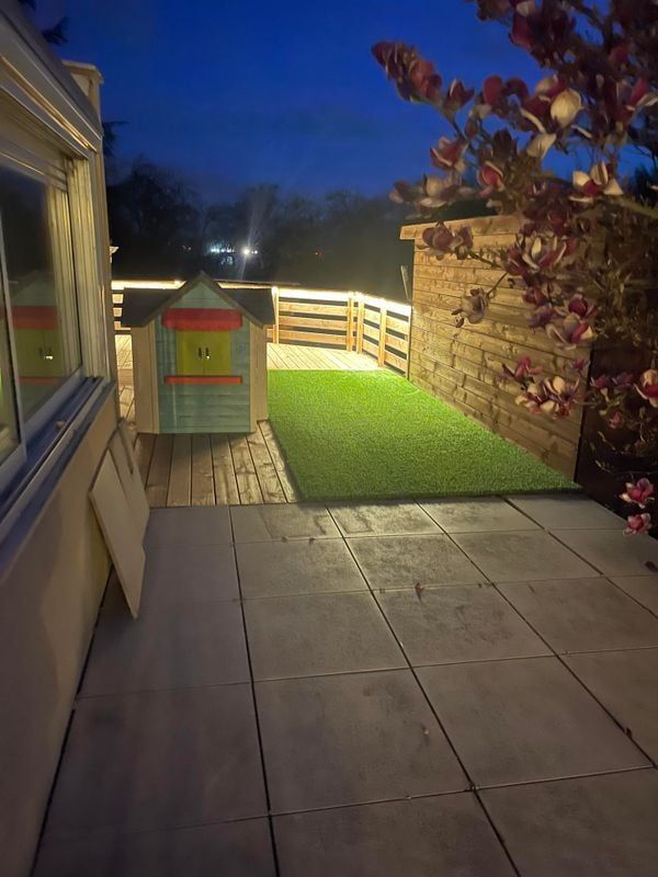Terrasse avec niche pour chien sur gazon artificiel, clôture en bois et arbre en fleurs, éclairée au crépuscule.