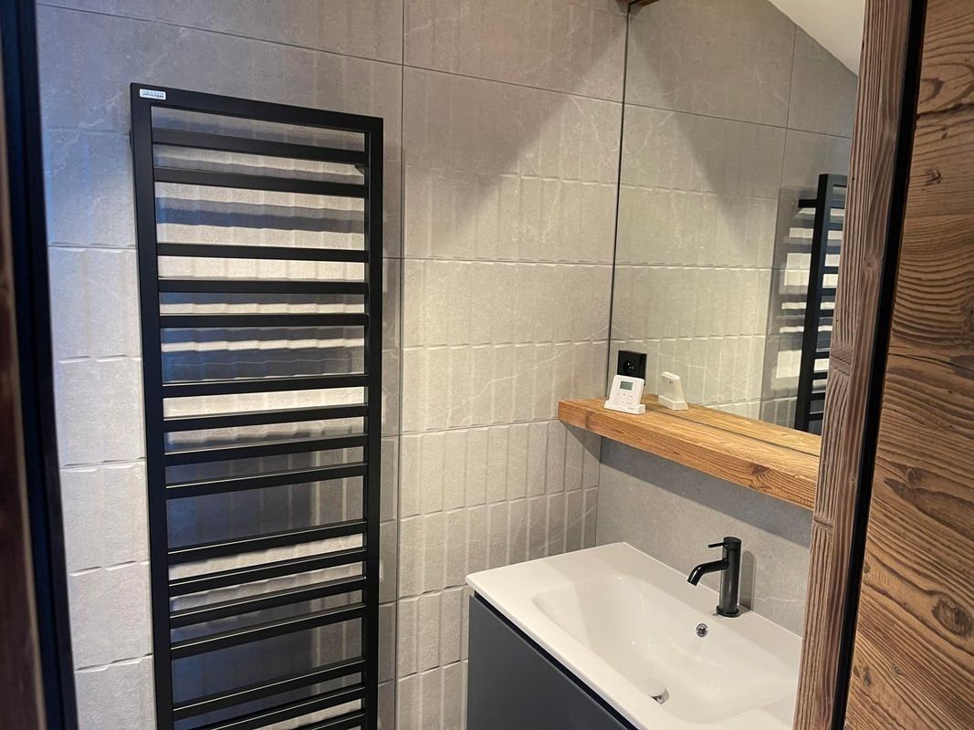 Salle de bains avec sèche-serviettes noir, carrelage gris, lavabo blanc et éléments en bois.