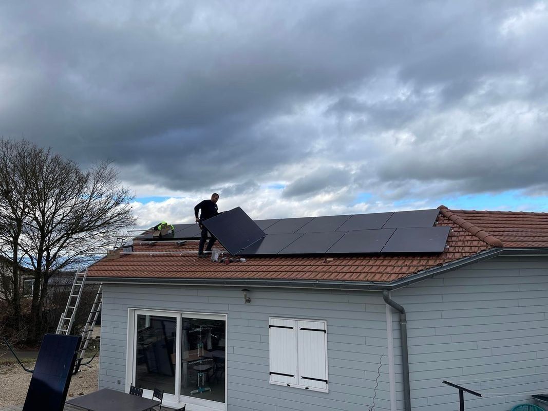 Un homme installe des panneaux solaires sur le toit d'une maison sous un ciel nuageux.