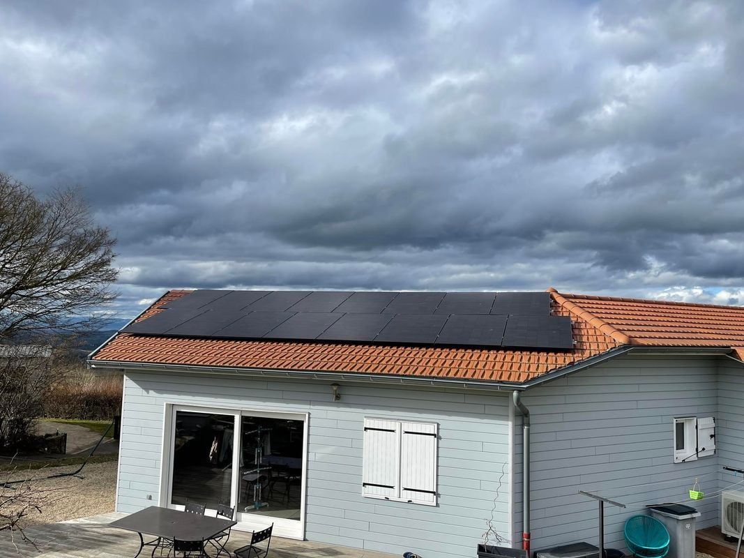 Panneaux solaires sur le toit en tuiles de terre cuite d'une maison grise, se détachant sur un ciel nuageux.