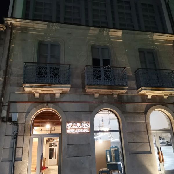 Fotografía nocturna de un edificio de tres plantas con puertas y balcones arqueados. Una tienda con un letrero luminoso: 
