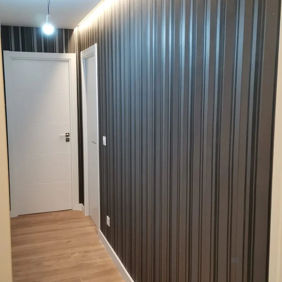 Pasillo estrecho con papel pintado oscuro con nervaduras verticales, puertas blancas y suelo de madera clara.