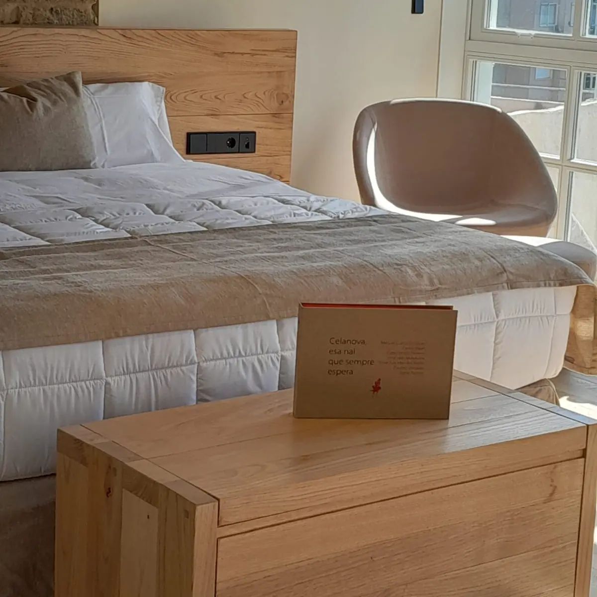 Dormitorio con cabecero de madera, sábanas de color beige, mesita de noche y una silla moderna cerca de una ventana.