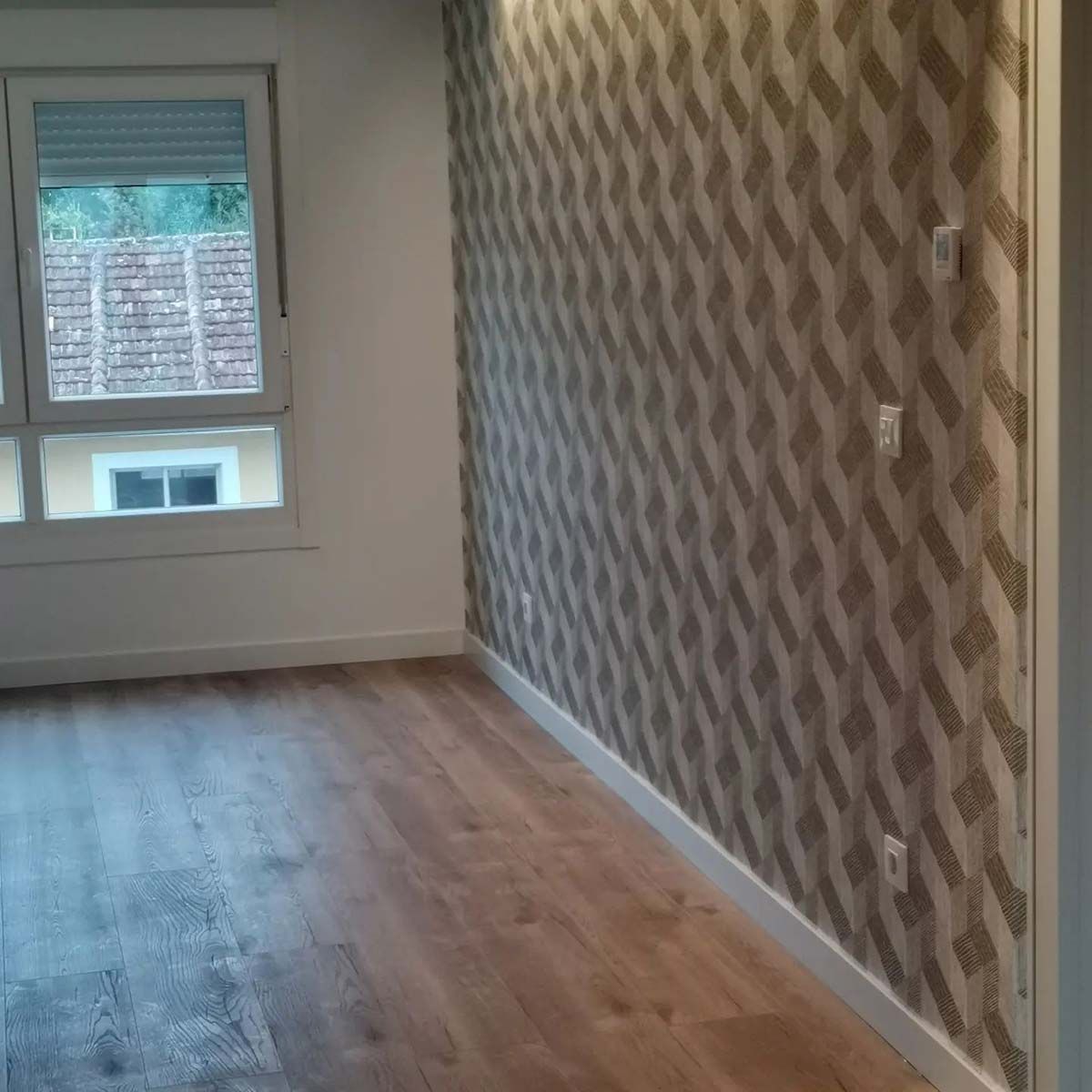 Habitación vacía con piso de madera, molduras blancas, ventana y papel tapiz estampado en la pared.