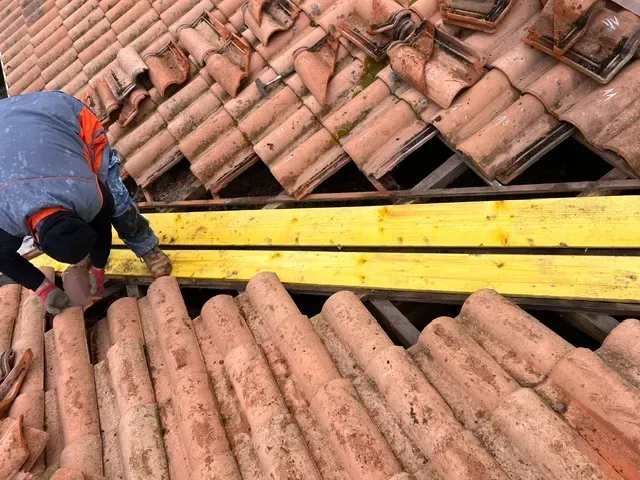 Un couvreur travaille sur un toit en tuiles de terre cuite, posant des planches jaunes.