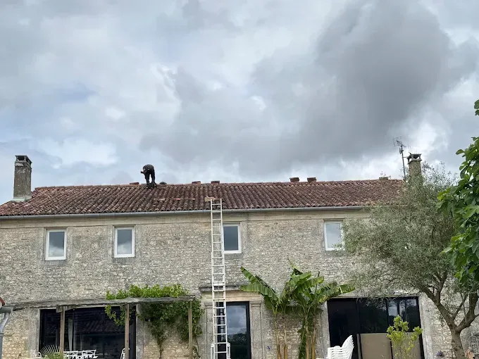 Une personne répare les tuiles d'une maison en pierre sur le toit, sous un ciel nuageux.
