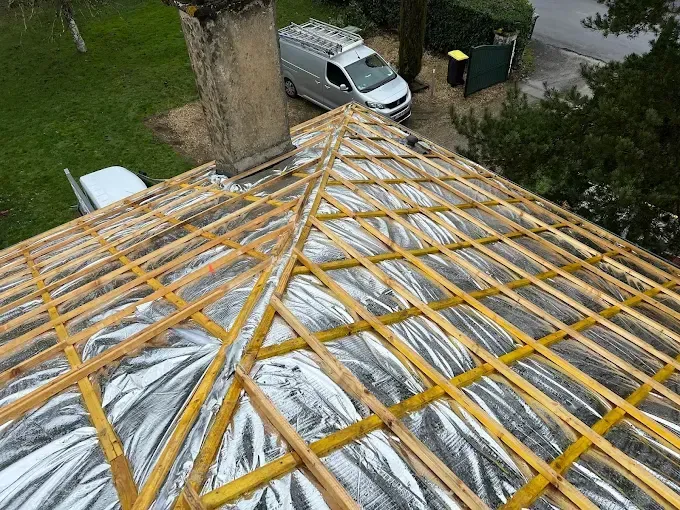 Charpente de toit avec isolation réfléchissante, cheminée et fourgonnette de chantier en arrière-plan.