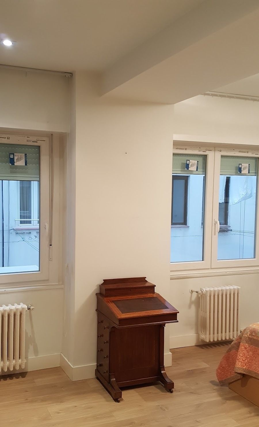 Habitación luminosa con dos ventanas, radiadores y un pequeño armario de madera en el suelo.