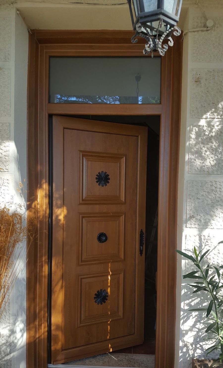 Puerta principal de madera con paneles decorativos y farolero, enmarcada por paredes de estuco color crema y una planta en maceta.