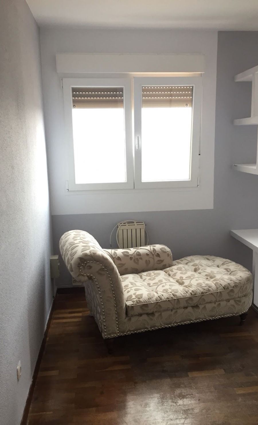Habitación pequeña y luminosa con una chaise longue blanca, ventana, radiador y suelo de madera oscura.