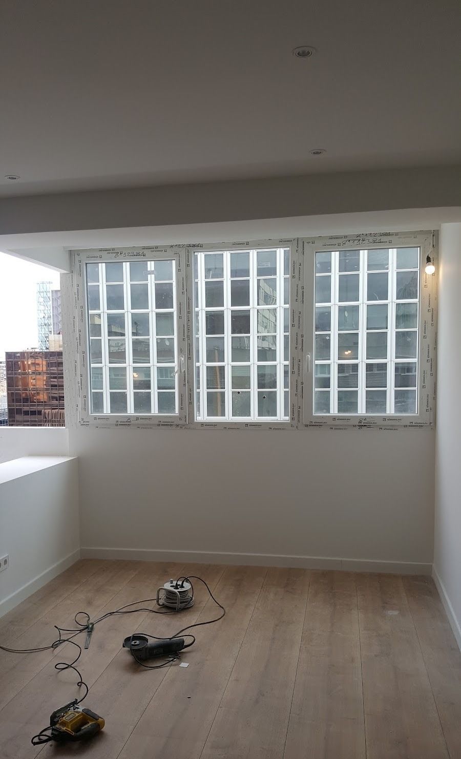Habitación vacía en obras con suelo de madera, herramientas esparcidas y grandes ventanales en la pared del fondo.