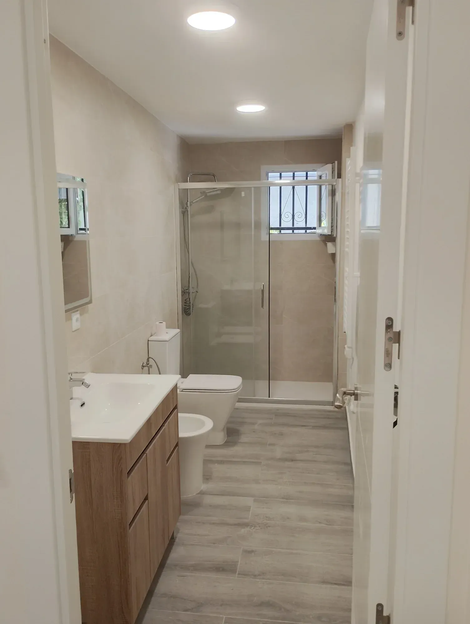 Baño moderno y estrecho, con mueble de madera, inodoro, ducha con mampara de vidrio y suelo gris.