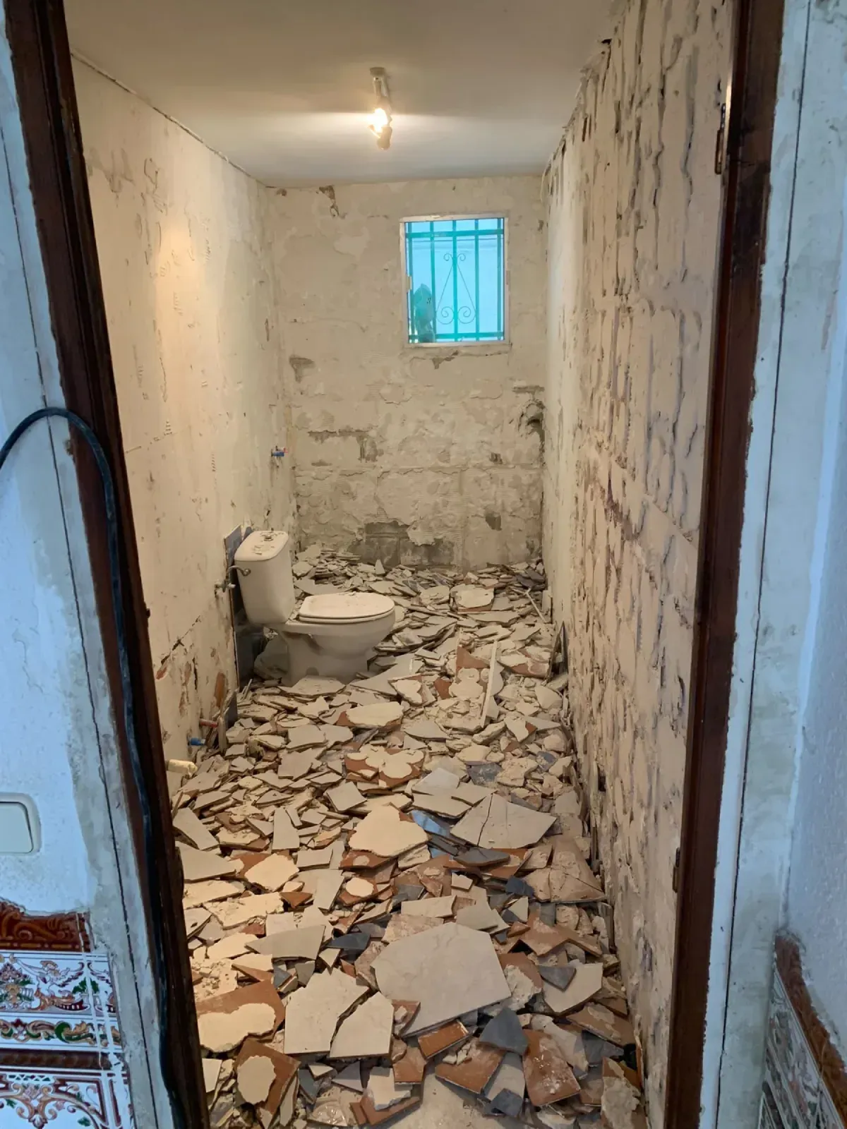 Baño dañado, con el suelo cubierto de escombros, paredes desconchadas, un inodoro y una pequeña ventana con barrotes