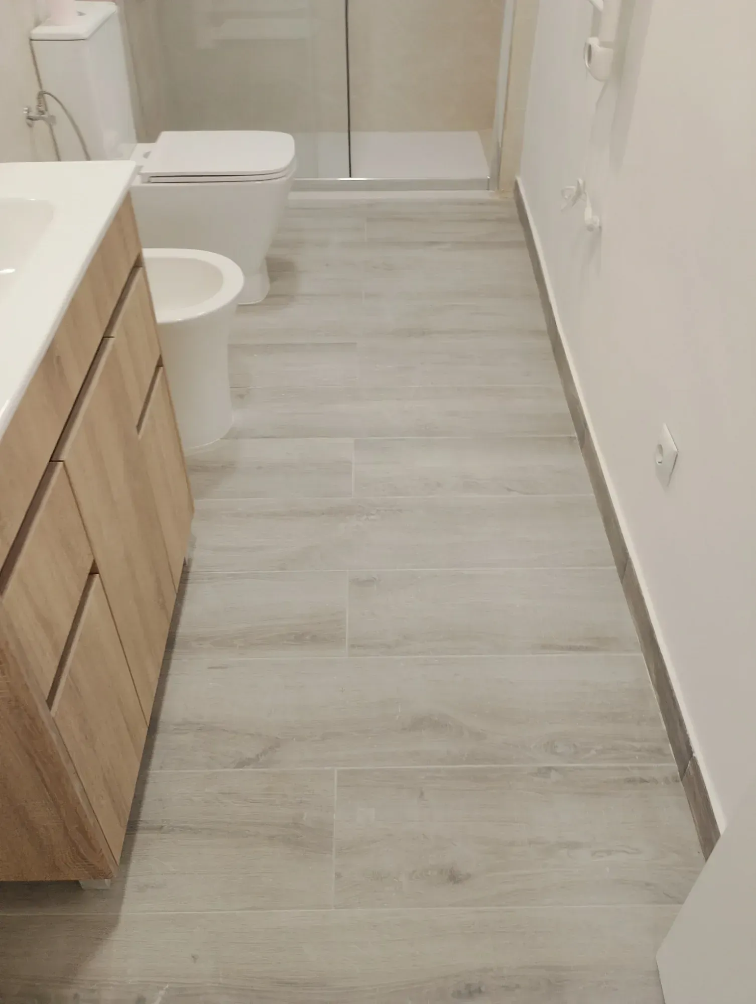 Baño estrecho con suelo gris efecto madera, paredes blancas, mueble de lavabo, inodoro y puerta de ducha.