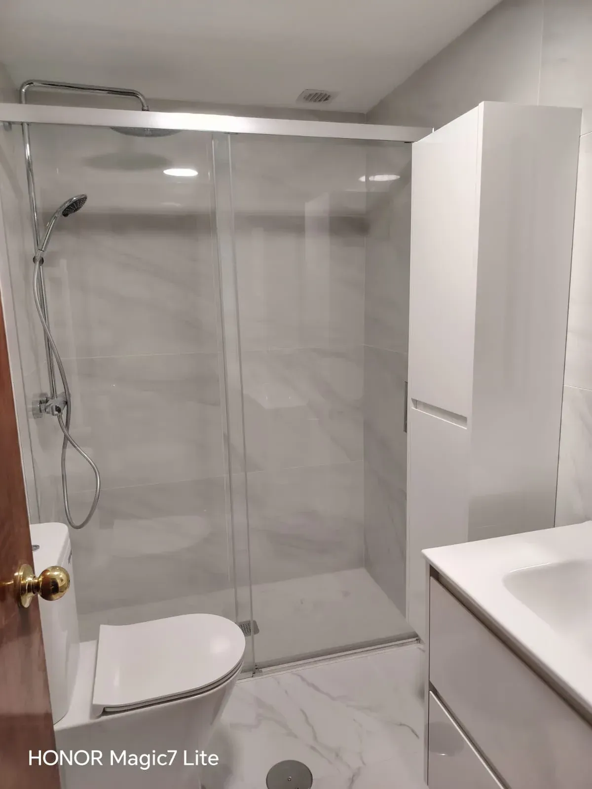 Baño moderno con mampara de ducha de vidrio, tocador blanco y suelo y paredes con diseño de mármol