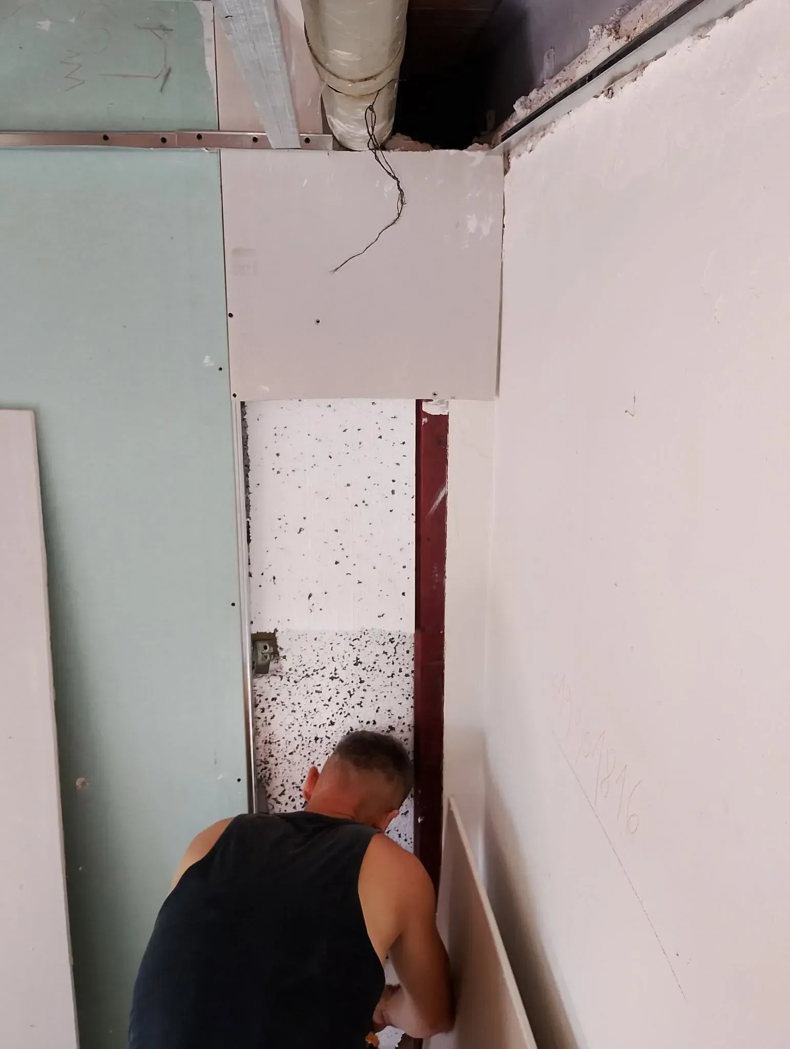 Una persona agachada en una puerta estrecha, trabajando cerca de una pared dañada con material del techo a la vista.
