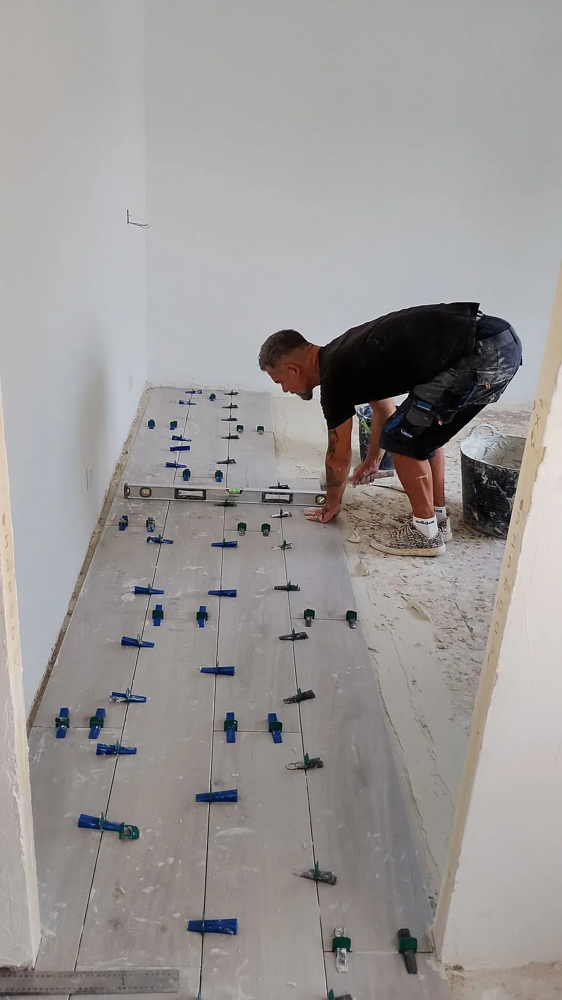 Un trabajador arrodillado sobre un suelo de baldosas, colocando baldosas con crucetas azules en una habitación blanca.