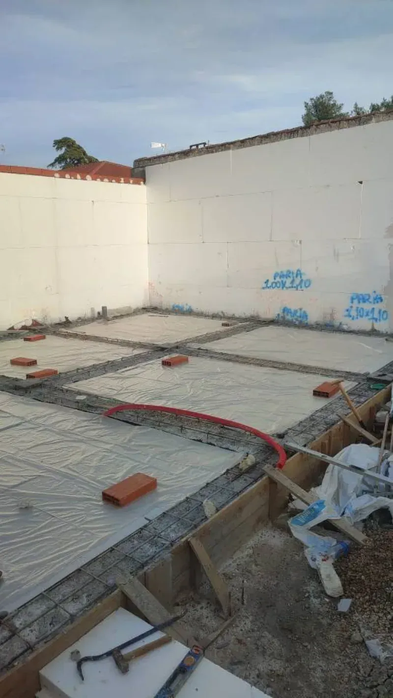 Obra con varillas de refuerzo y ladrillos dispuestos sobre una losa de hormigón junto a paredes blancas.