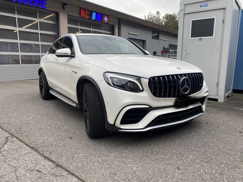 Une Mercedes Benz GLC 63 AMG blanche est garée devant un immeuble.