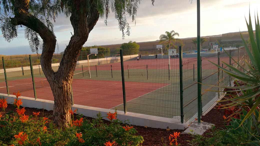 Una cancha de tenis con una valla alrededor y un árbol en primer plano.