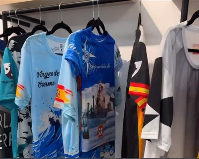 Camisetas con diseños estampados en azul, negro y blanco colgadas en un perchero. Algunas tienen imágenes de edificios y banderas.