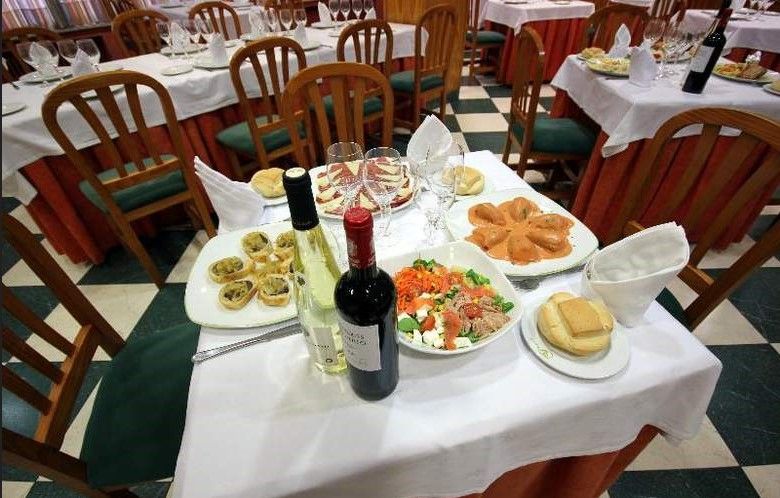 Mesa de restaurante preparada para comer, con platos de comida, botellas de vino y copas, rodeada de sillas y mesas.