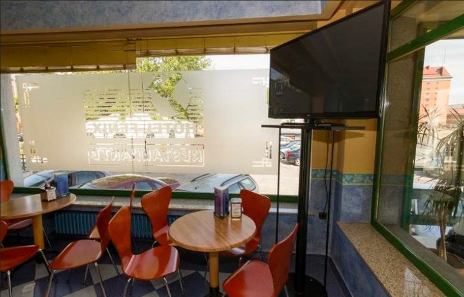 Interior de restaurante con mesas y sillas naranjas, un televisor grande y una ventana con diseño esmerilado.