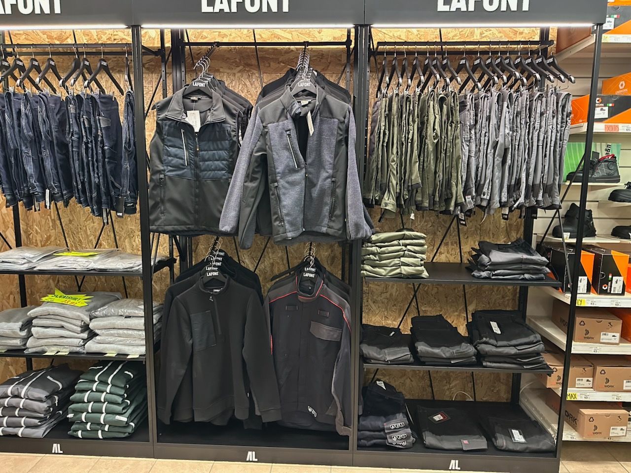 Présentoir de vêtements dans un magasin, comprenant des vestes, des chemises et des pantalons noirs, gris et verts.