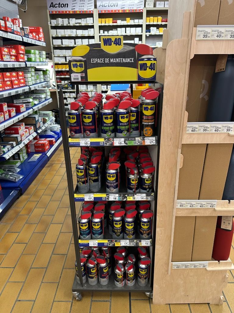 Présentation de bombes aérosol WD-40 sur une étagère de magasin, étiquettes rouges et jaunes, fond d'allée de magasin.