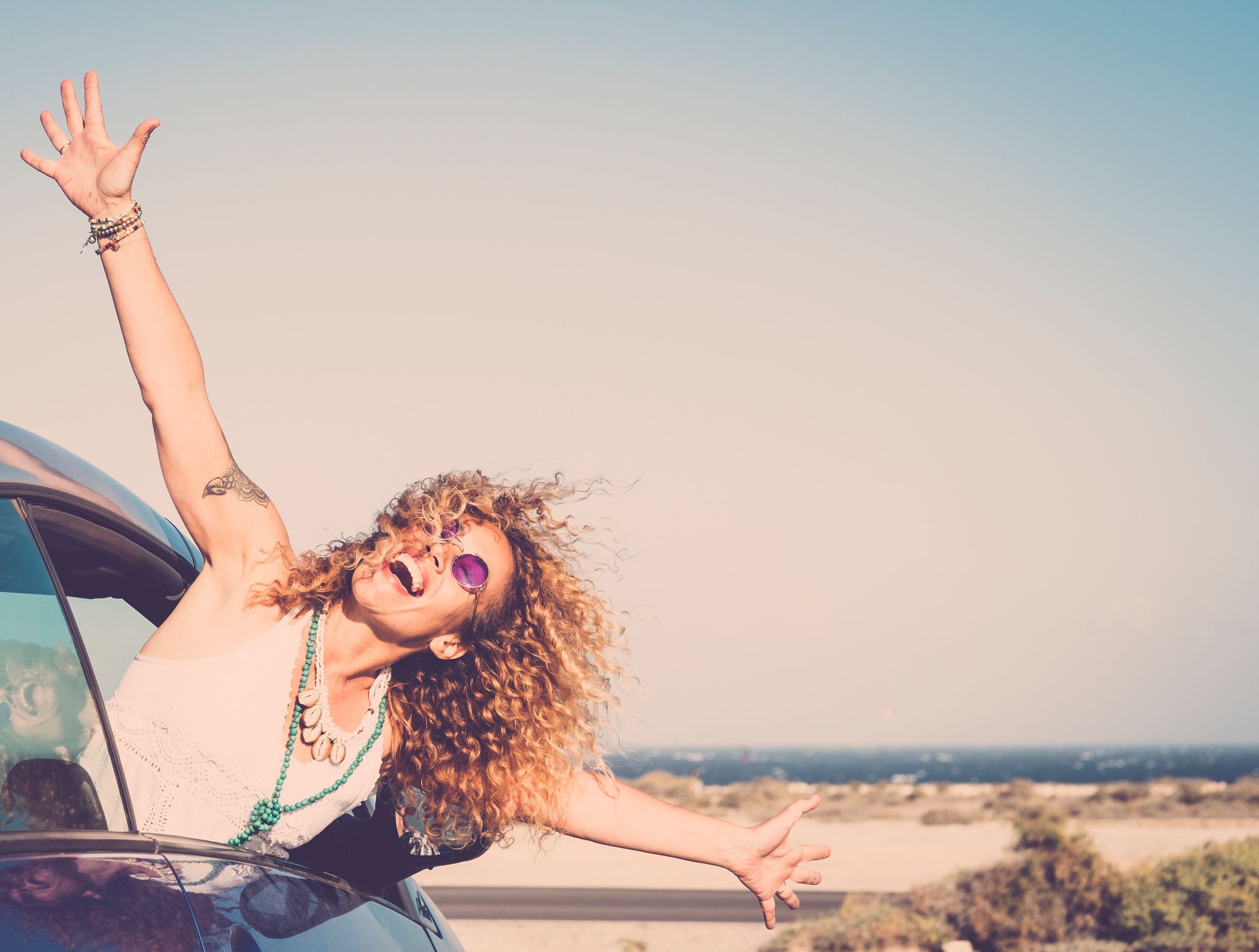 Woman with curly hair leans out of car window, arms raised in joy at the beach.
