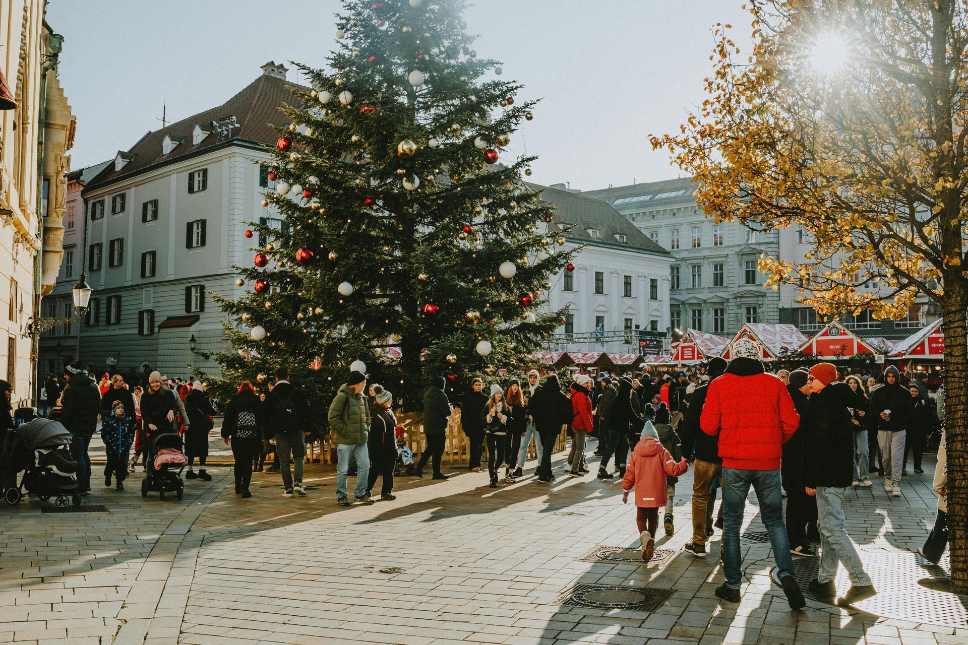 Christmas market with a decorated tree, bustling crowds, and sunny cityscape.