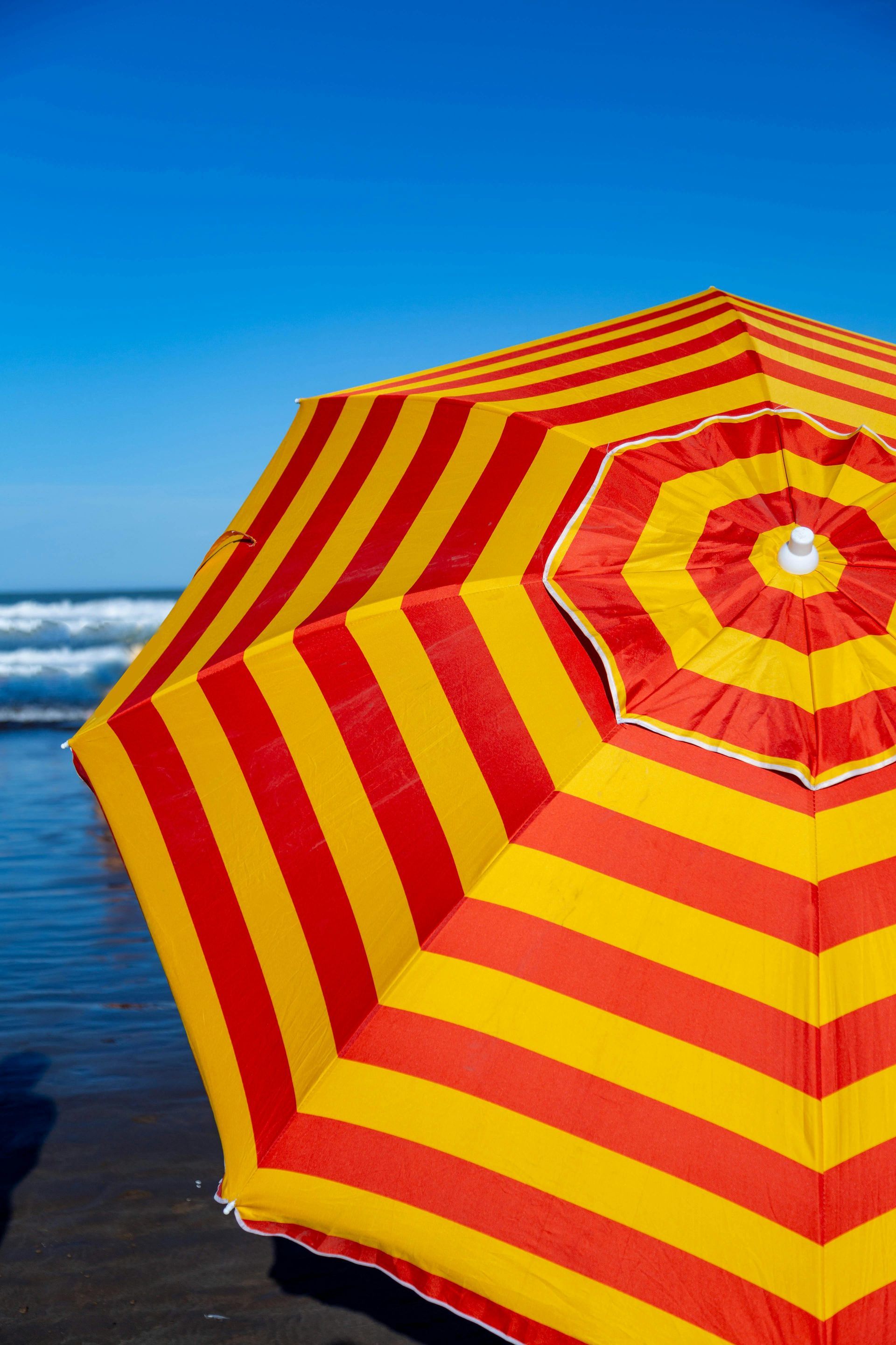 Striped red and yellow beach umbrella on a dark sand beach with waves and blue sky.