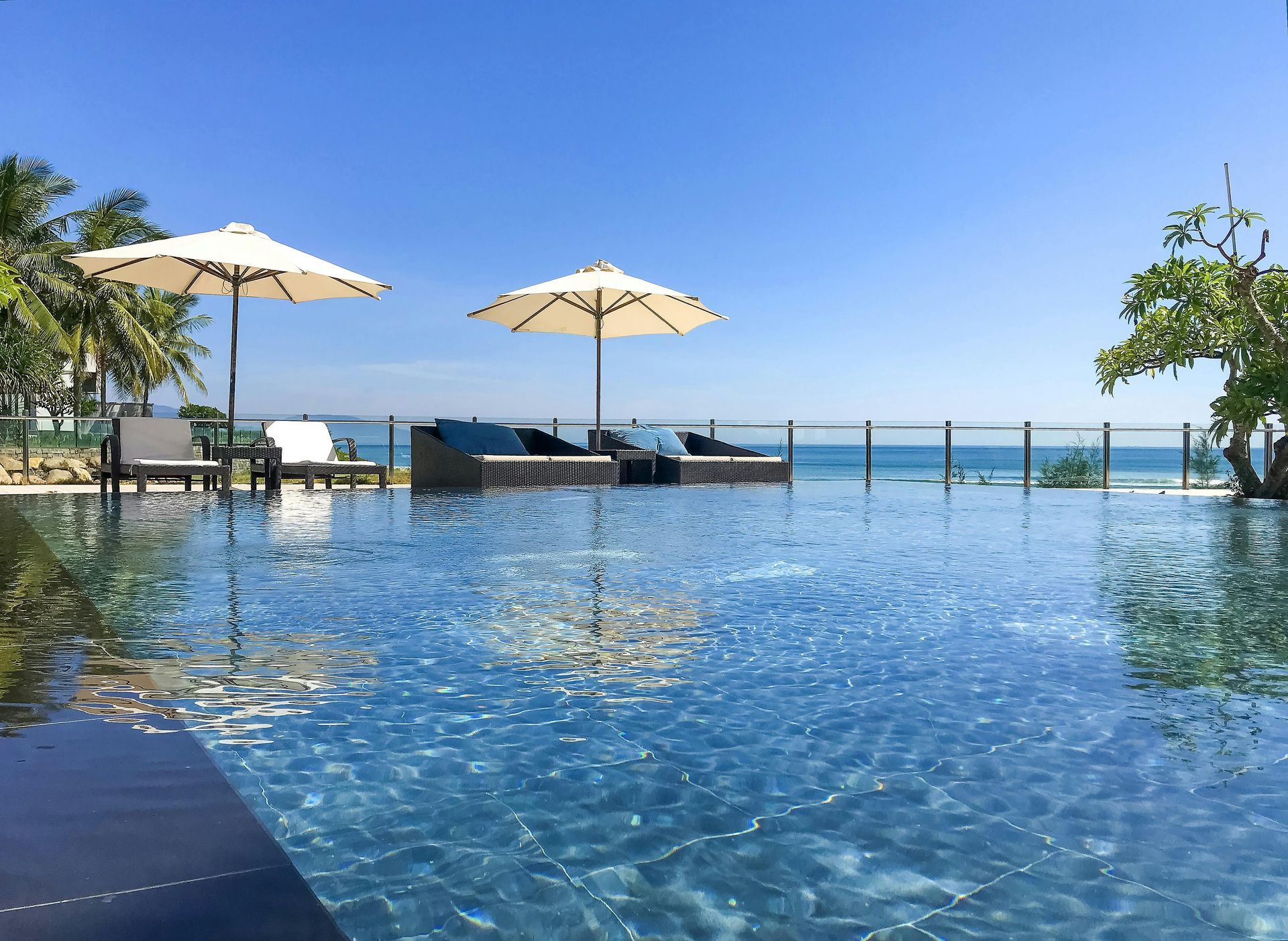 Swimming pool overlooking ocean with umbrellas and lounge chairs under a clear blue sky.