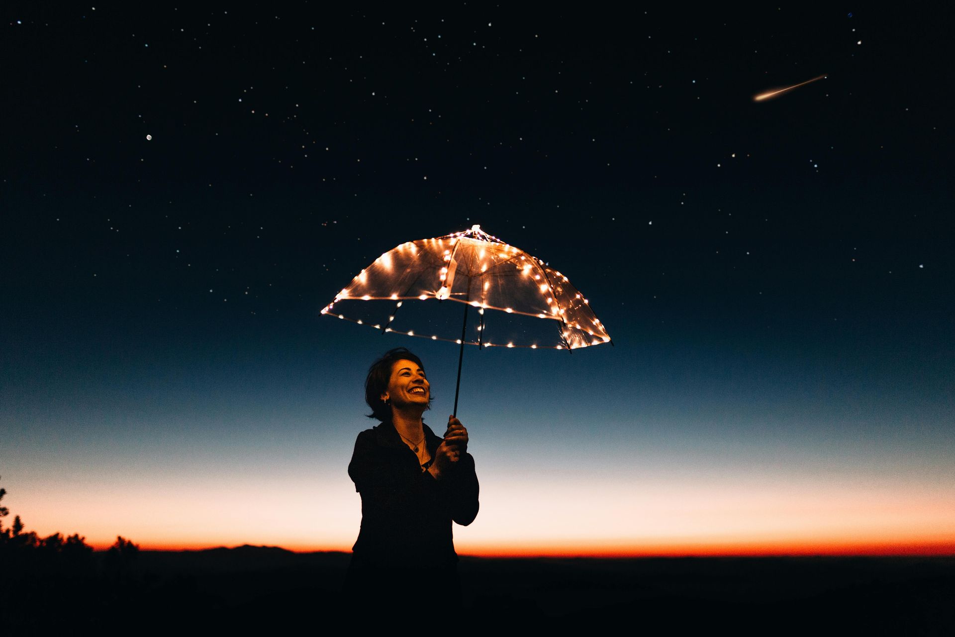 Woman holding lit umbrella, looking up at night sky with shooting star. Silhouette against orange and blue sunset.